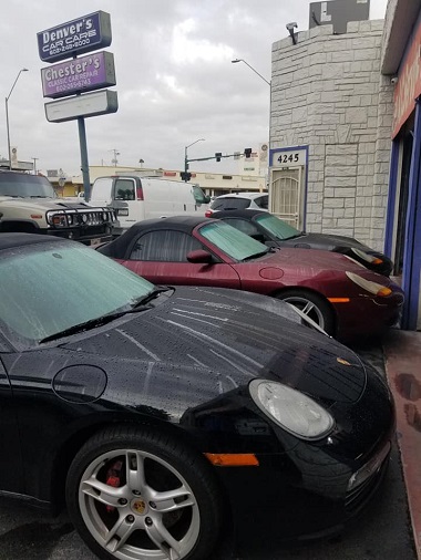 Three Porsche Boxster cars in the parking lot at Denver's Car Care in Phoenix, Arizona. The foremost car has is all black, followed by a red and black car, and finally, another all black Boxter. The sign for Denver's Care Care and Chester's Classic Car Repair can be seen in the background.